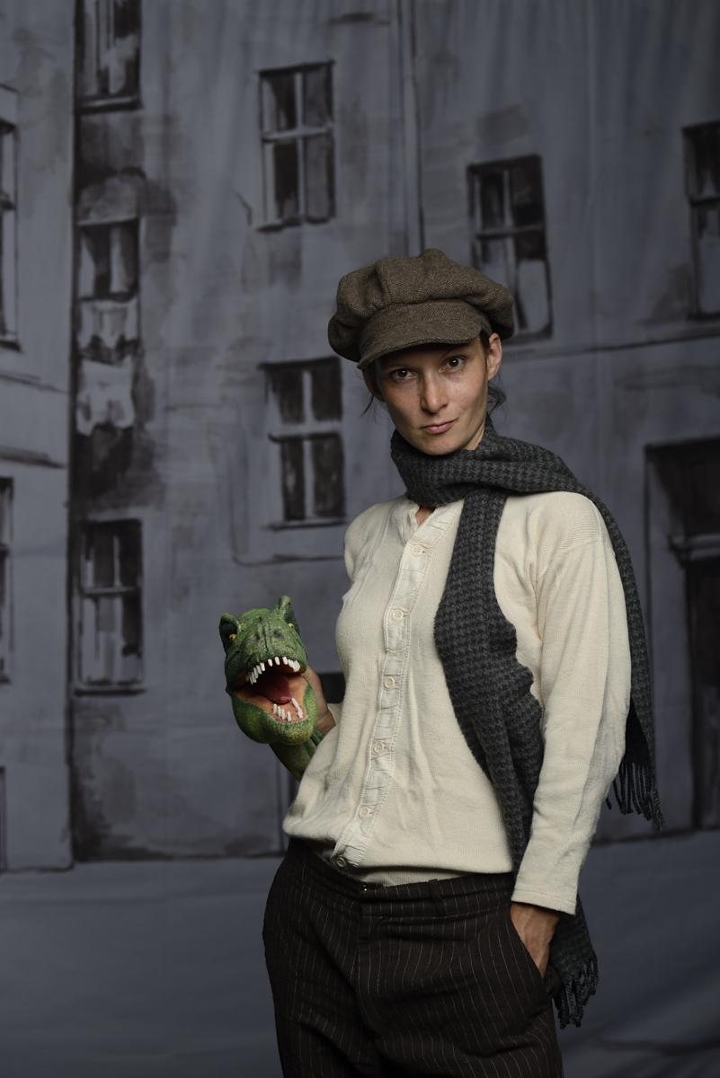 Die Puppenspielerin steht vor einer grauen Häuserwand. Sie hält einen grünen Drachen in der Hand