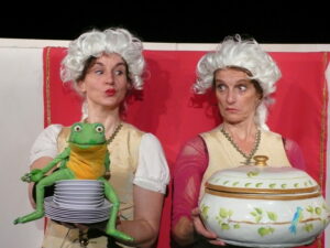zwei Spielerinnen mit weißen Perücken halten einen Frosch und eine große Suppenschüssel hoch. Sie machen spitze Münder.