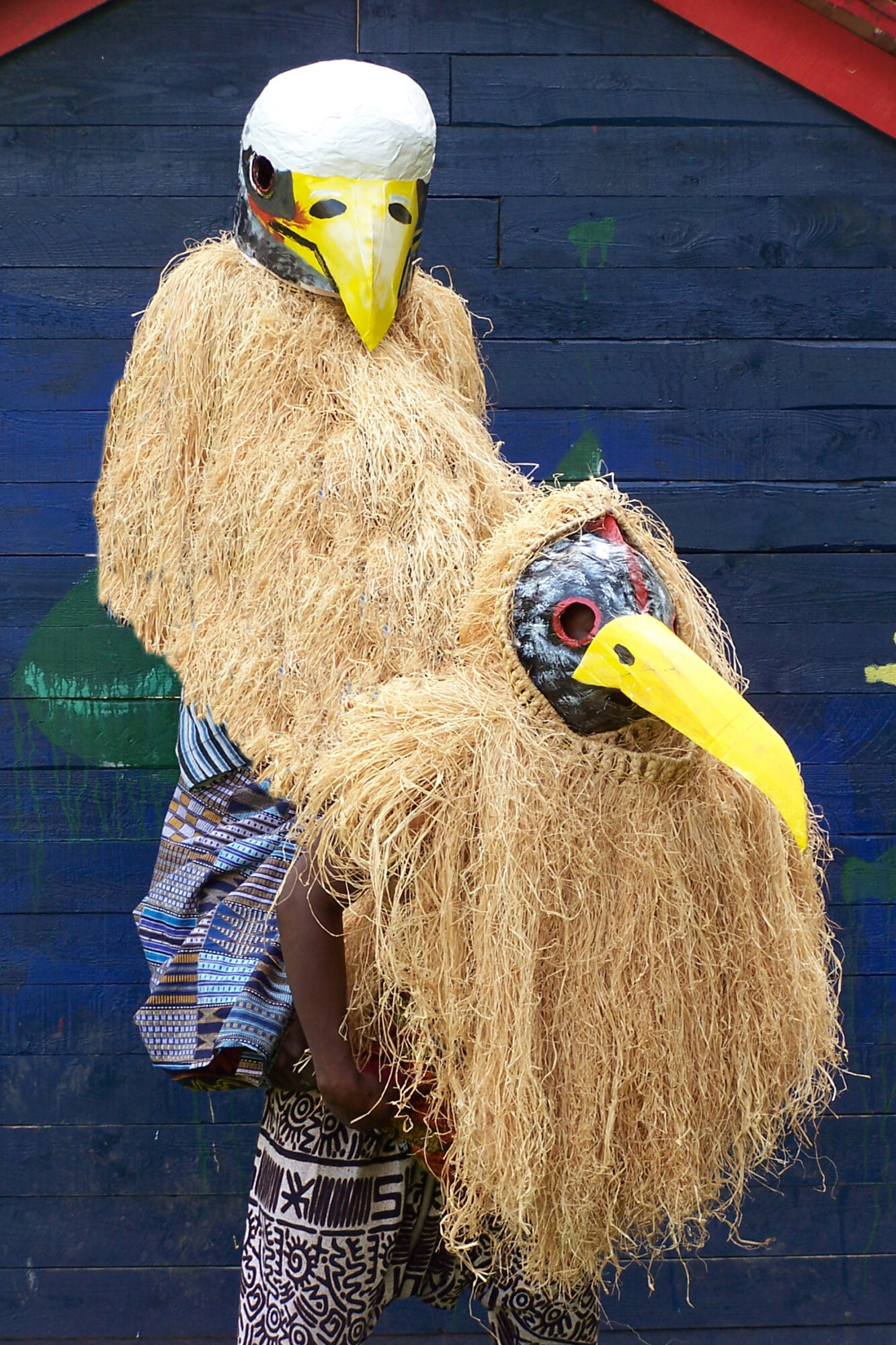 zwei Künstler von Adesa sind mit Masken als Vögel verkleidet. Sie stehen vor einer blauen Wand.