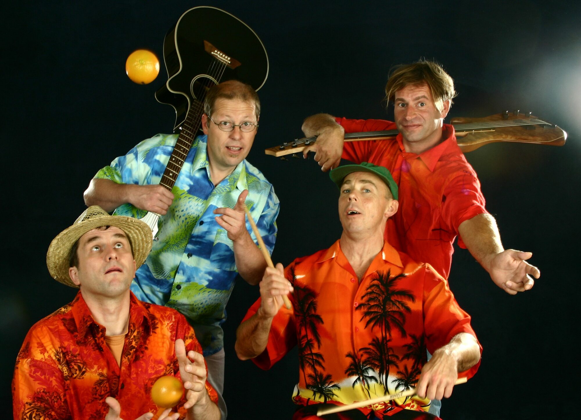 4 Musiker stehen vor einem schwarzen Hintergrund. Sie tragen Hawai-Hemden und haben Instrumente in der Hand. Einer wirft eine Orange